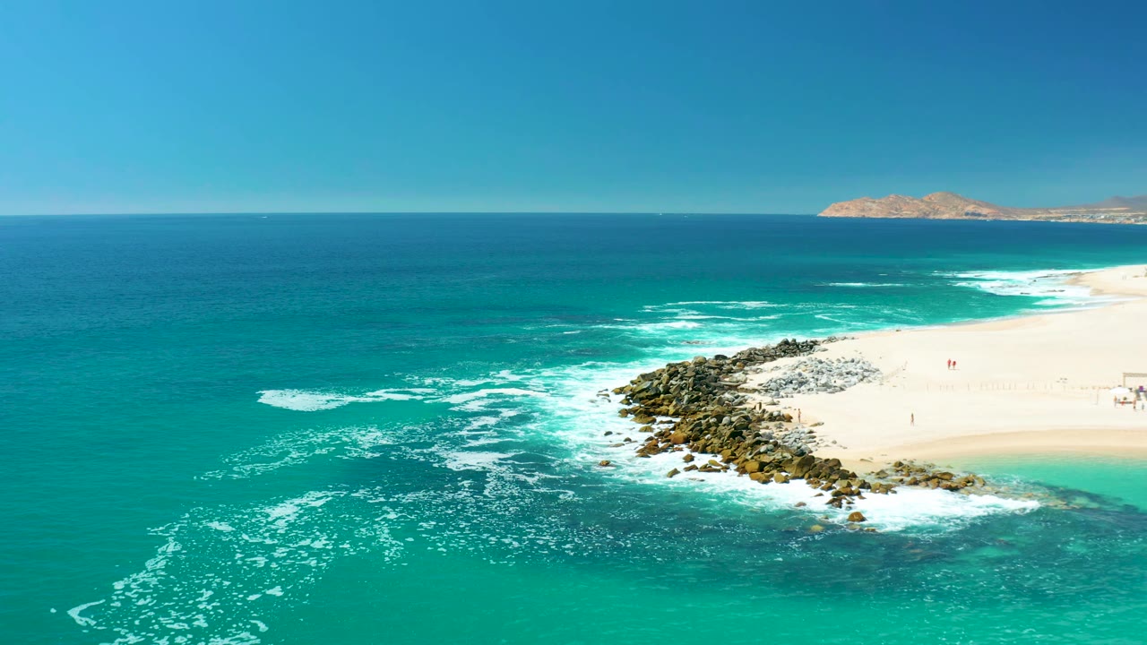 Turquoise surf wraps around a rocky point with sand and a distant island beyond.