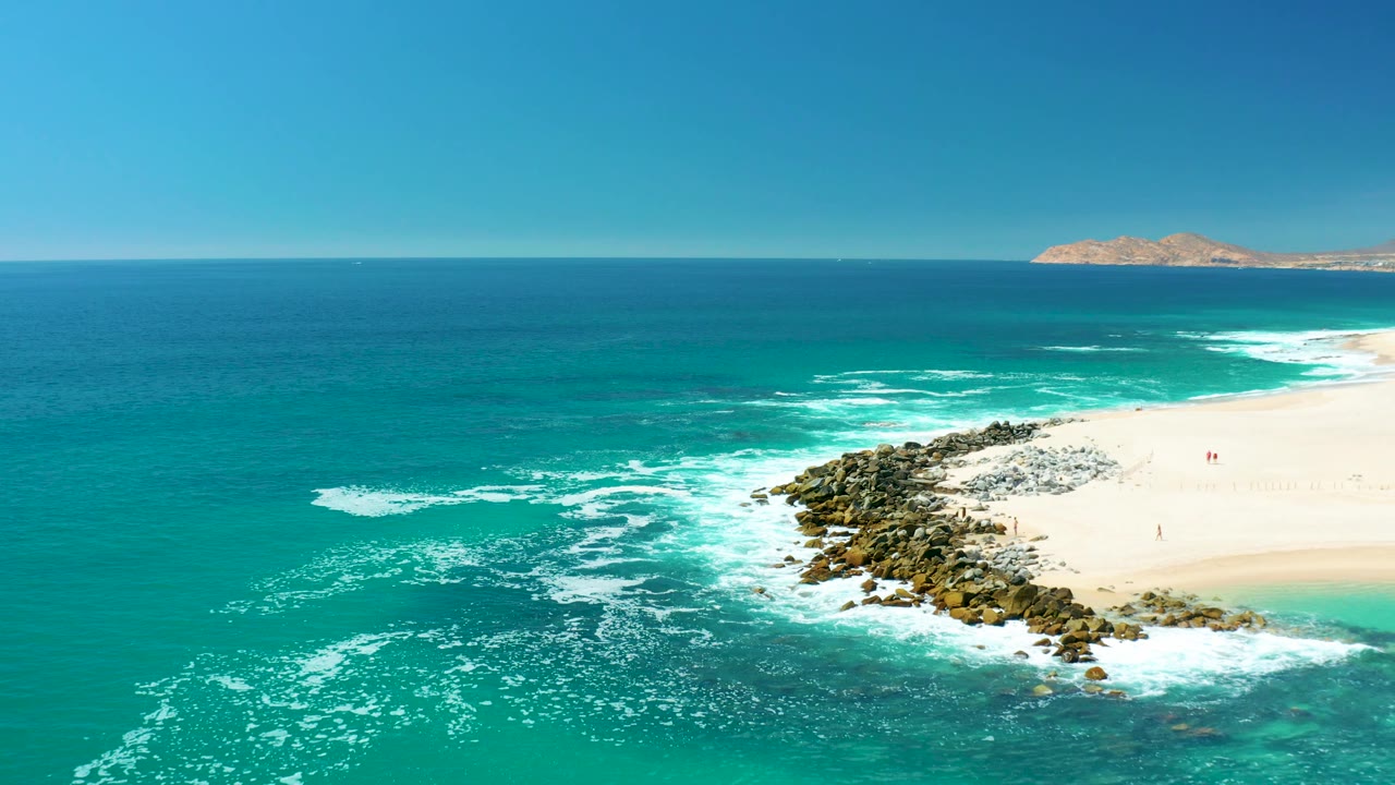 The frame shows Marquis Los Cabos surf pushing around the rocks, with more beach visible at right, open turquoise water at left, and a low island across the clear horizon.