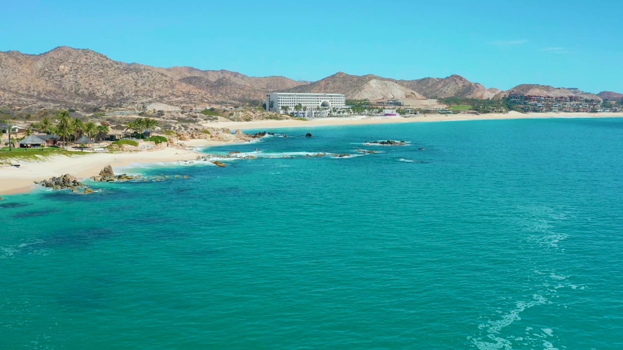 Wide aerial frame at Marquis Los Cabos in Mexico showing blue-green water across the foreground, small white foam trails near shore, beach rocks on the left, distant buildings, mountains, and no visible people.