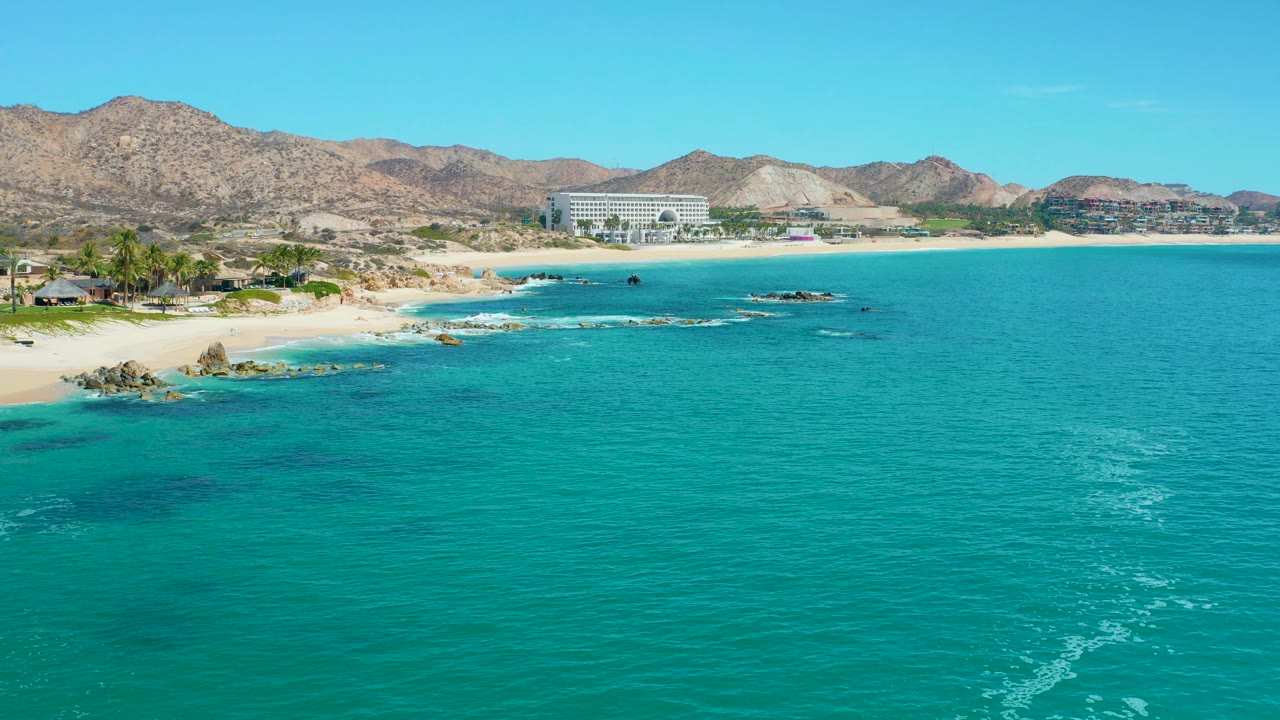 Ocean foreground with white foam lines, sandy beach, rocks, buildings, and mountain ridge.
