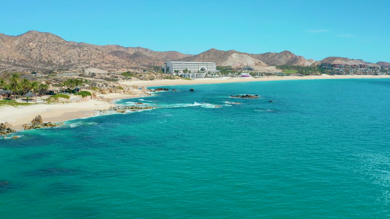 Opening aerial frame of Marquis Los Cabos, Mexico, with turquoise water in front, a sandy beach and dark rocks at left, resort buildings near the center, distant dry mountains, and tiny beachgoers visible.