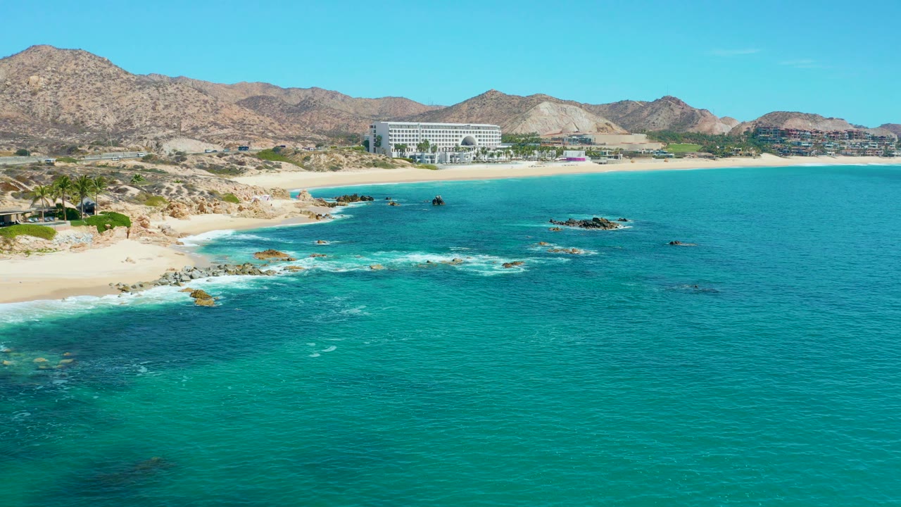 At 0:00, a wide elevated view at Marquis Los Cabos shows turquoise water in the foreground, waves breaking on rocks, a sandy beach at left, a large coastal building, mountains, and no visible people.