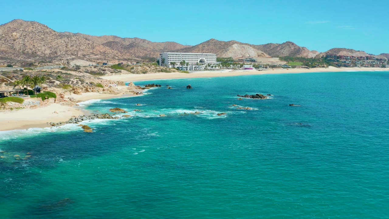 At 0:01, the Marquis Los Cabos shoreline remains in a wide aerial view as white foam spreads around the offshore rocks, with the sandy beach, large building, mountains, and no visible people behind it.