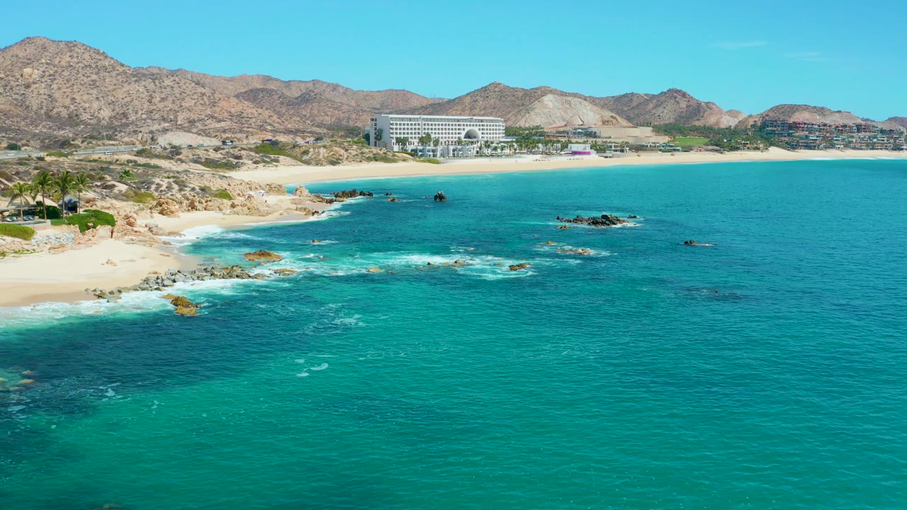 At 0:02, the wide view at Marquis Los Cabos shows more white water along the left shoreline rocks, turquoise ocean filling the foreground, the beach and large building in the distance, and no visible people.