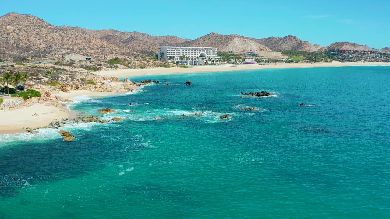 Wide coastal aerial with turquoise sea, rocky surf, sandy beach, large building, and dry mountains.