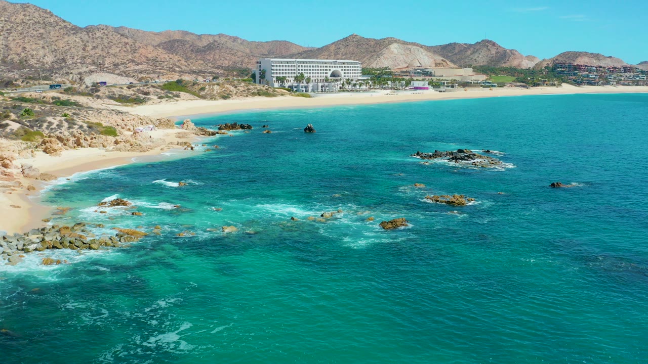 Aerial frame from Marquis Los Cabos, Mexico showing turquoise water and white foam around rock outcrops, with a sandy beach, large white building, mountains, and no visible people.