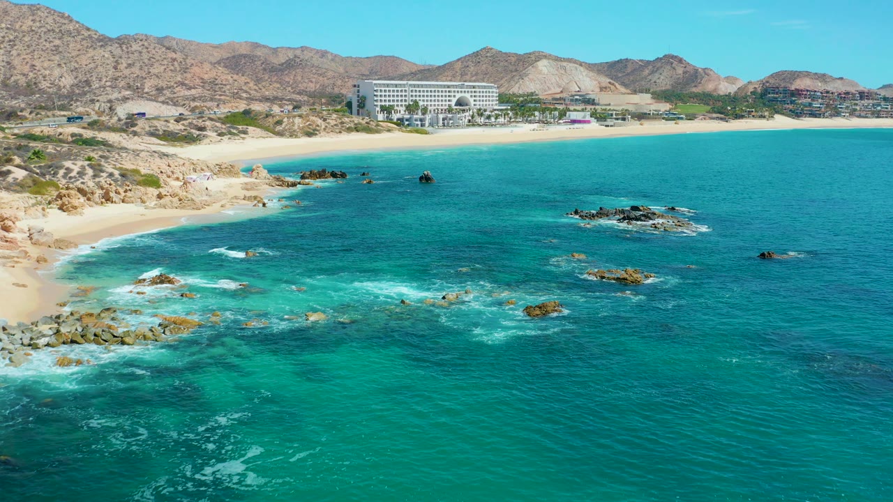 Aerial coastal view with turquoise water, surf on rocks, sandy beach, resort building, and mountains.