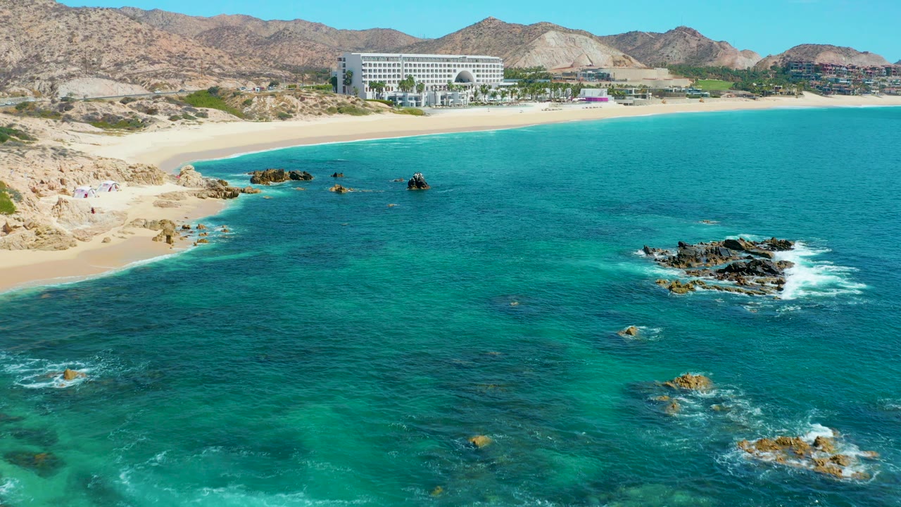 Aerial frame over Marquis Los Cabos showing turquoise water in the foreground, rocky outcrops with white foam, pale beach, a large shoreline building, mountain ridges, and no visible people.