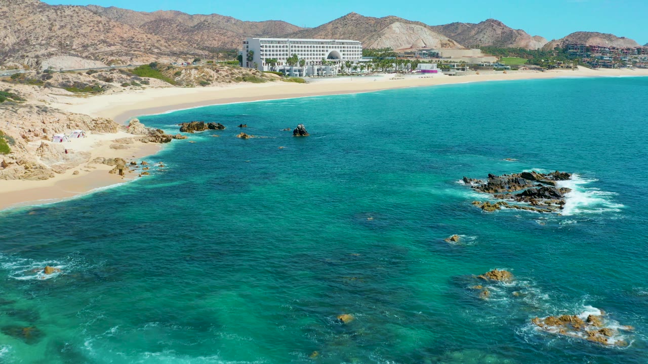 At 0:01, the Marquis Los Cabos aerial view keeps the beach and large shoreline building high in frame while small white wave breaks form around the darker rocks, with no visible people.