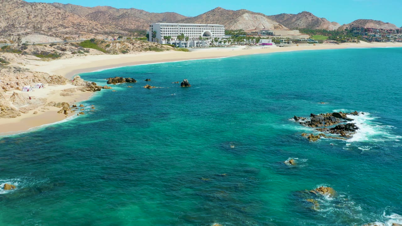 At 0:02, the drone view shows more open turquoise water across the center, with foaming rocks to the right, the sandy Marquis Los Cabos beach and large building above, and no visible people.