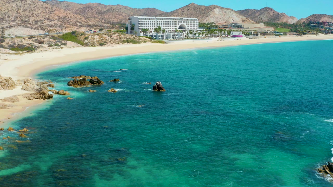 Aerial view of turquoise ocean, sandy beach, rocks, hotel, and mountains.