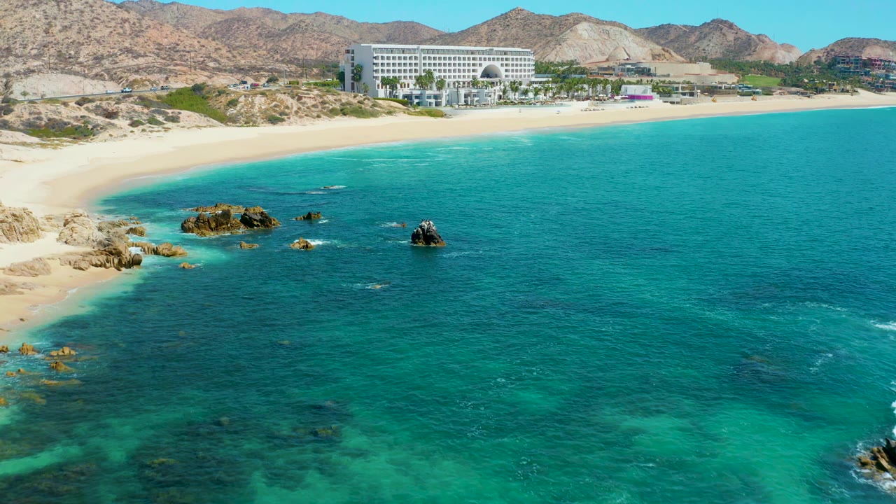 At 0:01, the Marquis Los Cabos shoreline remains wide in frame, with shallow turquoise water, scattered rocks, pale sand, a hotel building, mountains, and no visible people.