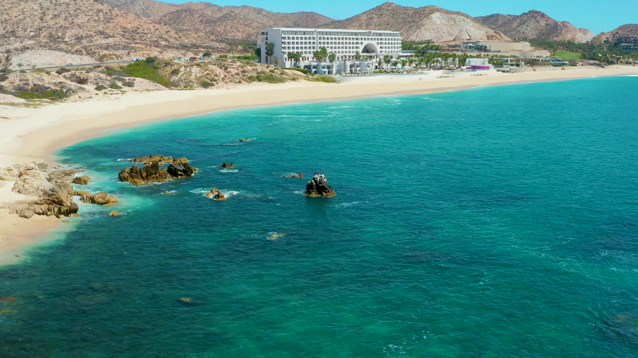 At 0:03, Marquis Los Cabos appears behind the curving sand while turquoise water and dark rock formations occupy the lower frame, with no visible people on the beach or in the water.