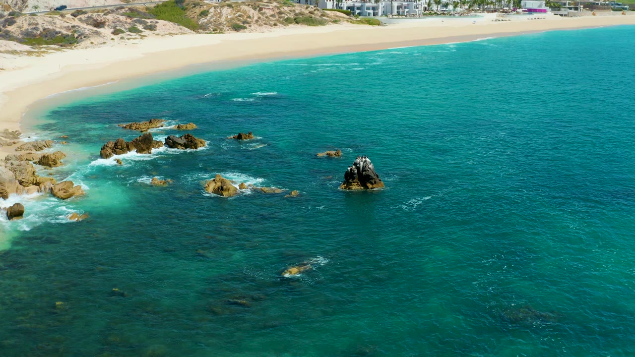 Aerial frame at Marquis Los Cabos, Mexico showing turquoise water around scattered rocks, a sandy beach along the top edge, and no visible people.