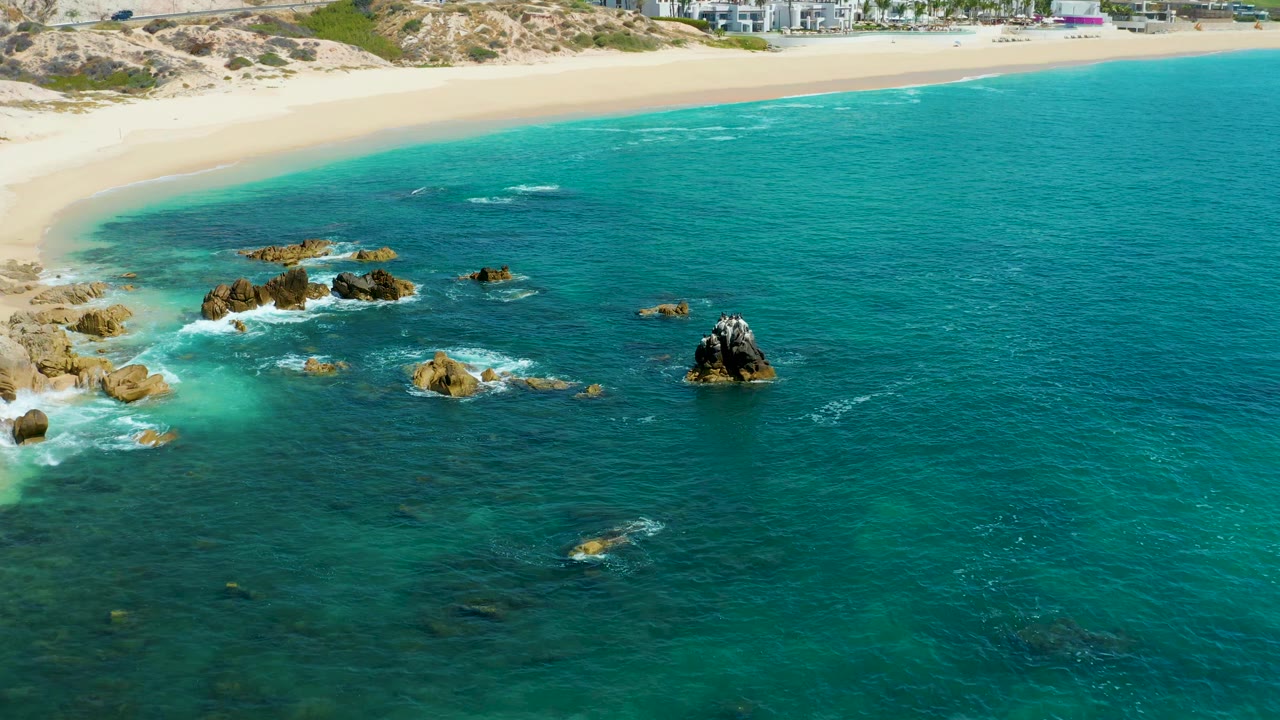 Aerial view at Marquis Los Cabos, Mexico with the sandy shoreline high in frame, dark and tan rocks in shallow turquoise water, and no visible people.