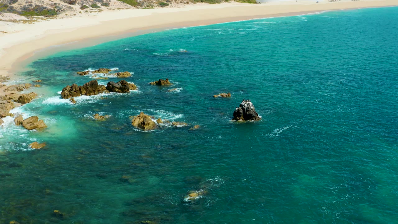 Ocean waves break around several rocks below a sandy shore.