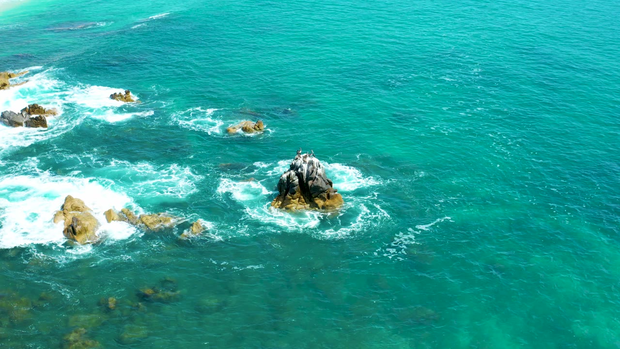 Aerial frame from Marquis Los Cabos, Mexico, showing foamy wave wash spreading farther around the left rock group and the central dark outcrop, no visible people.
