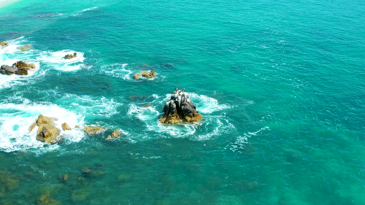 Aerial view over Marquis Los Cabos, Mexico, where the central rock remains ringed by white foam and open turquoise water fills the upper frame, no visible people.