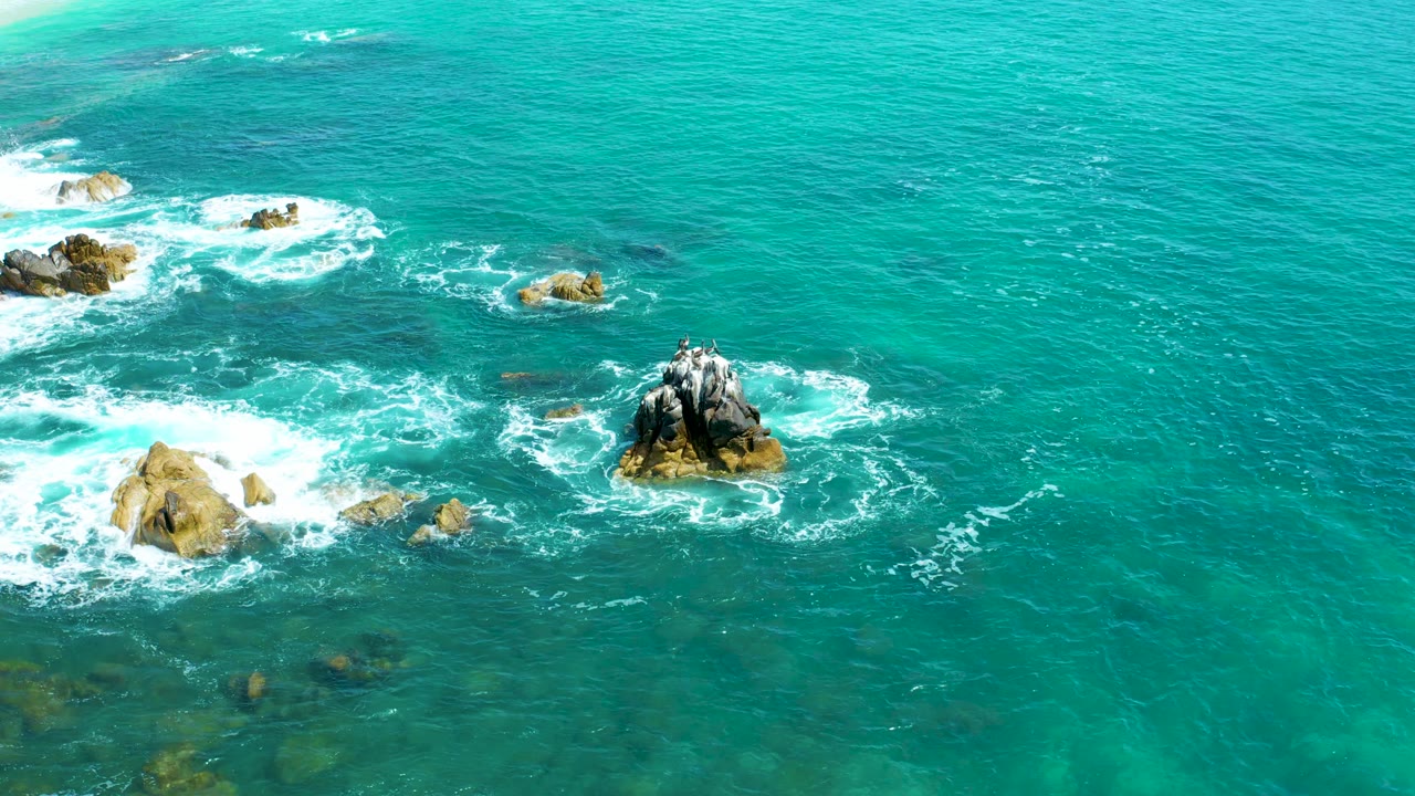 Aerial frame at Marquis Los Cabos, Mexico, with a larger tan rock near the lower left, a dark rock outcrop to the right, and fresh white foam between them, no visible people.