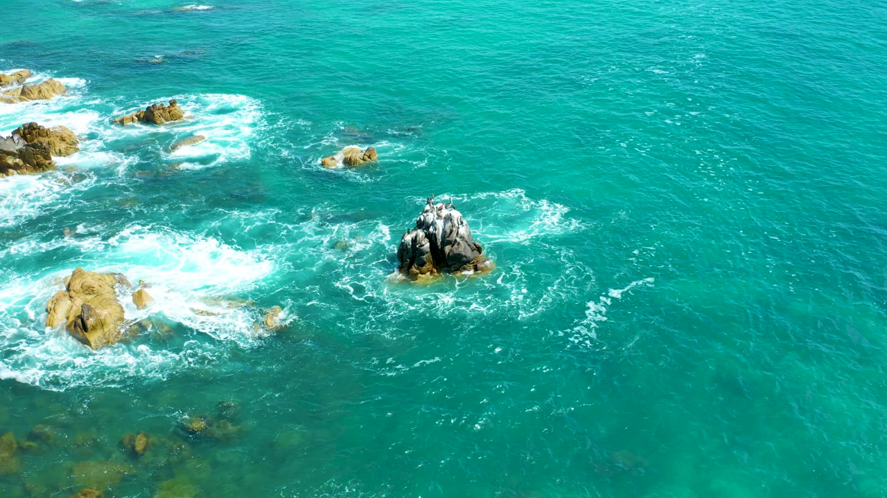 A central dark rock formation sits in bright turquoise water at Marquis Los Cabos while white surf curls around nearby tan rocks, with no visible people.