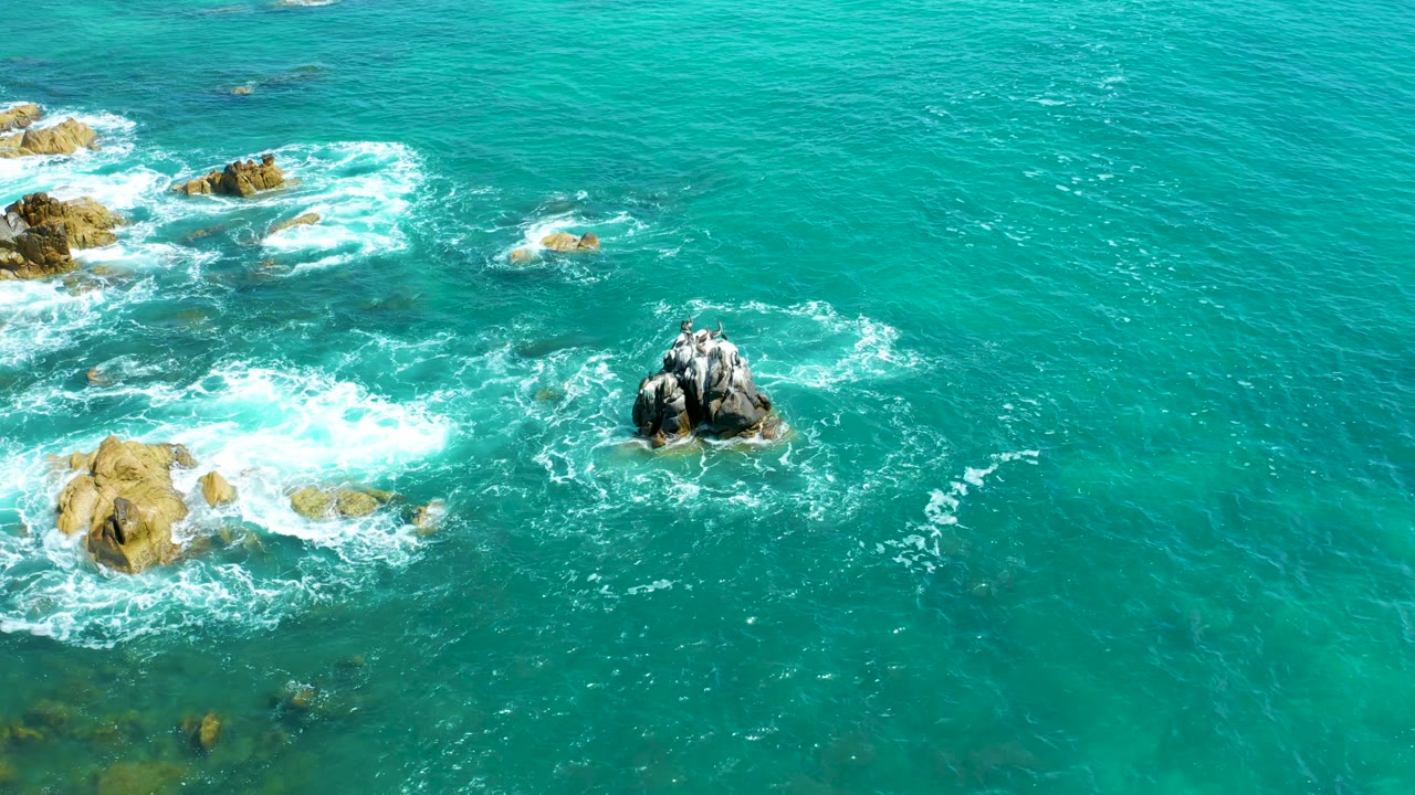 From above, white water spreads over the left rock cluster while a darker central outcrop remains surrounded by turquoise sea at Marquis Los Cabos, with no visible people.