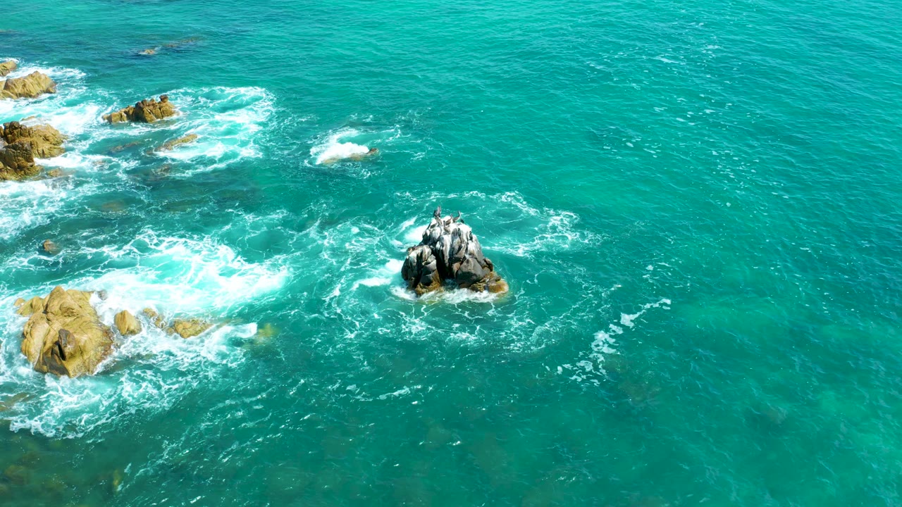 White foam pushes across the lower-left surf zone as the main rock outcrop remains right of center in turquoise water at Marquis Los Cabos, with no visible people.
