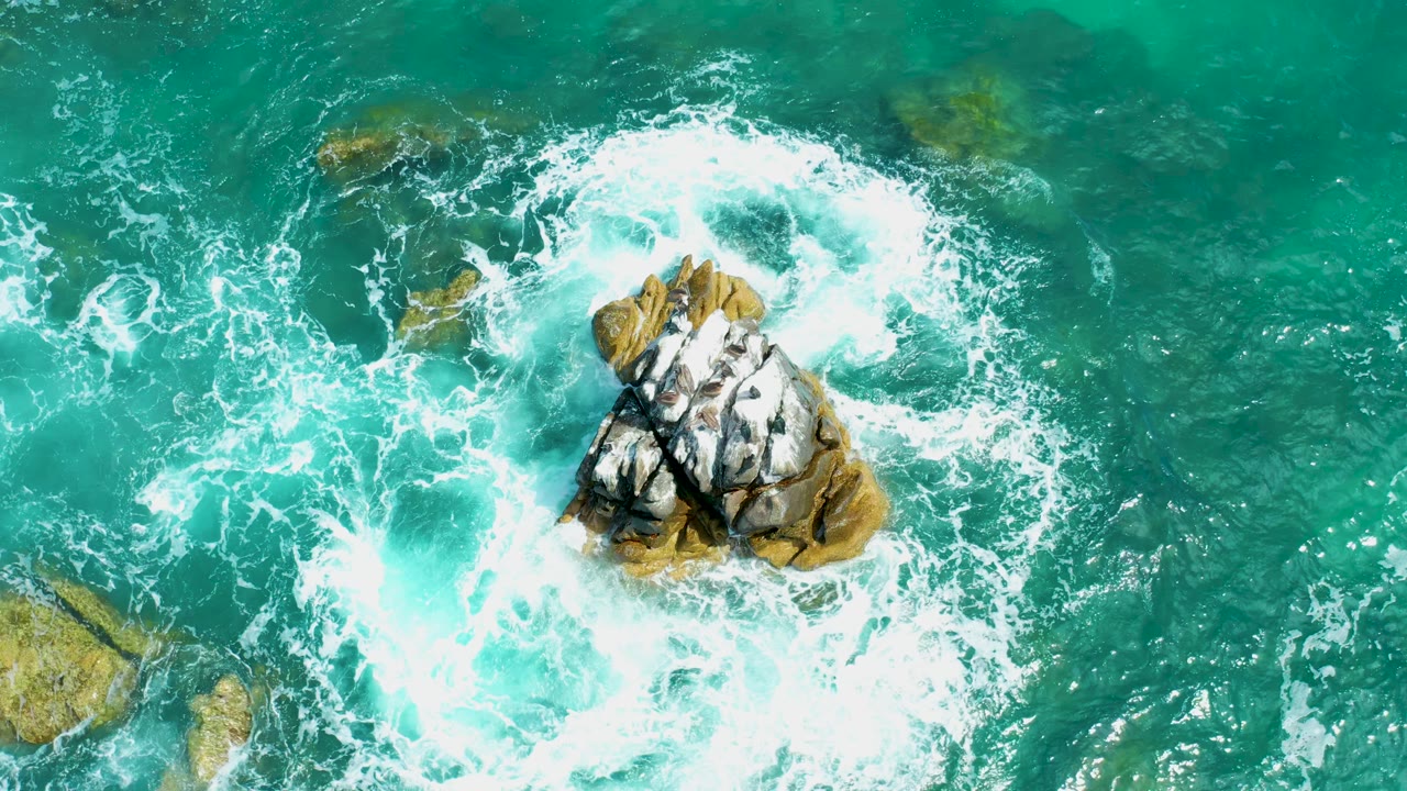 Aerial frame from Marquis Los Cabos, Mexico with foamy whitewater expanding around the central rock and clear green-blue water surrounding it, with no visible people.