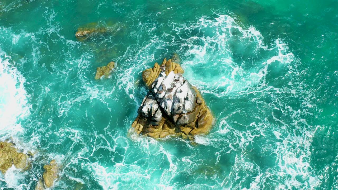 Overhead view at Marquis Los Cabos, Mexico of turquoise water swirling around a central rocky outcrop, with white foam forming to the left and no visible people.
