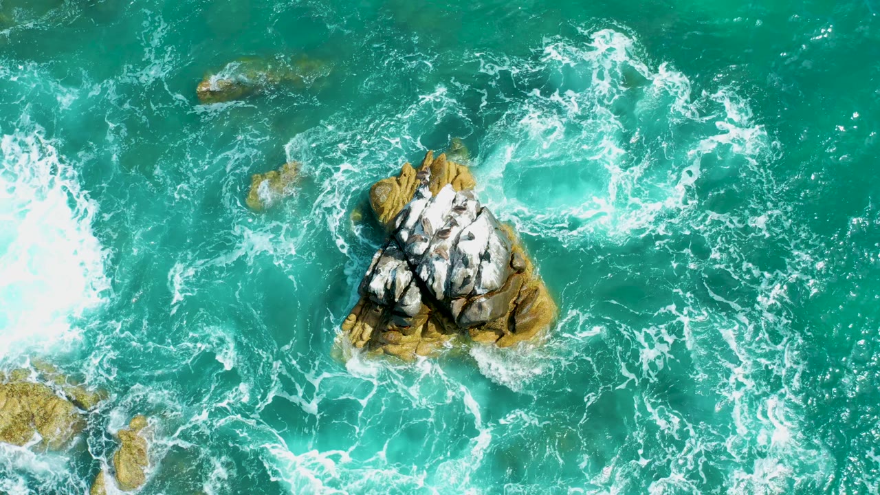Overhead view at Marquis Los Cabos, Mexico with bright white foam spreading across turquoise water beside the central rock outcrop, with no visible people.