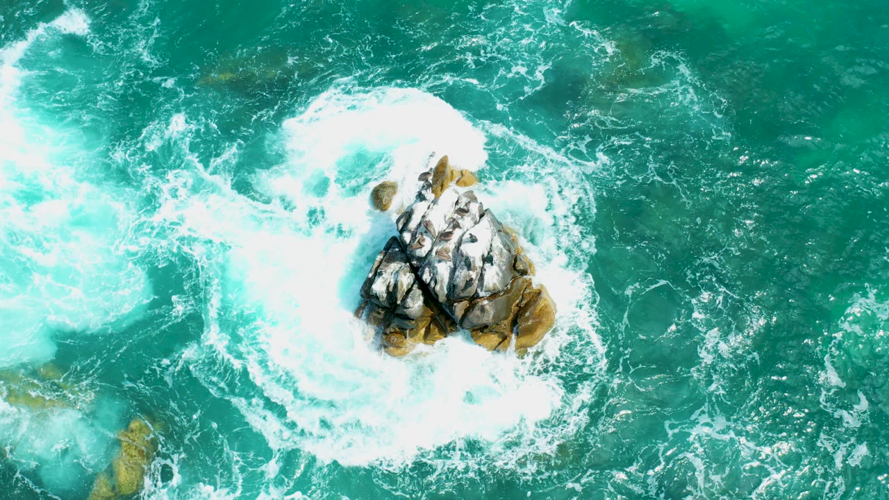 Overhead view at Marquis Los Cabos, Mexico, where bright white surf wraps around a tan and gray rocky outcrop in turquoise water, with no visible people.