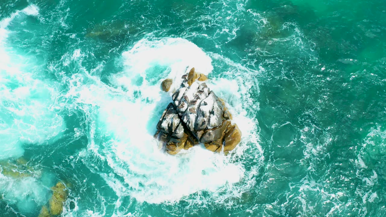 Aerial frame at Marquis Los Cabos, Mexico, showing foamy white water expanding left of the central rocky outcrop while deeper turquoise water fills the right side, with no visible people.