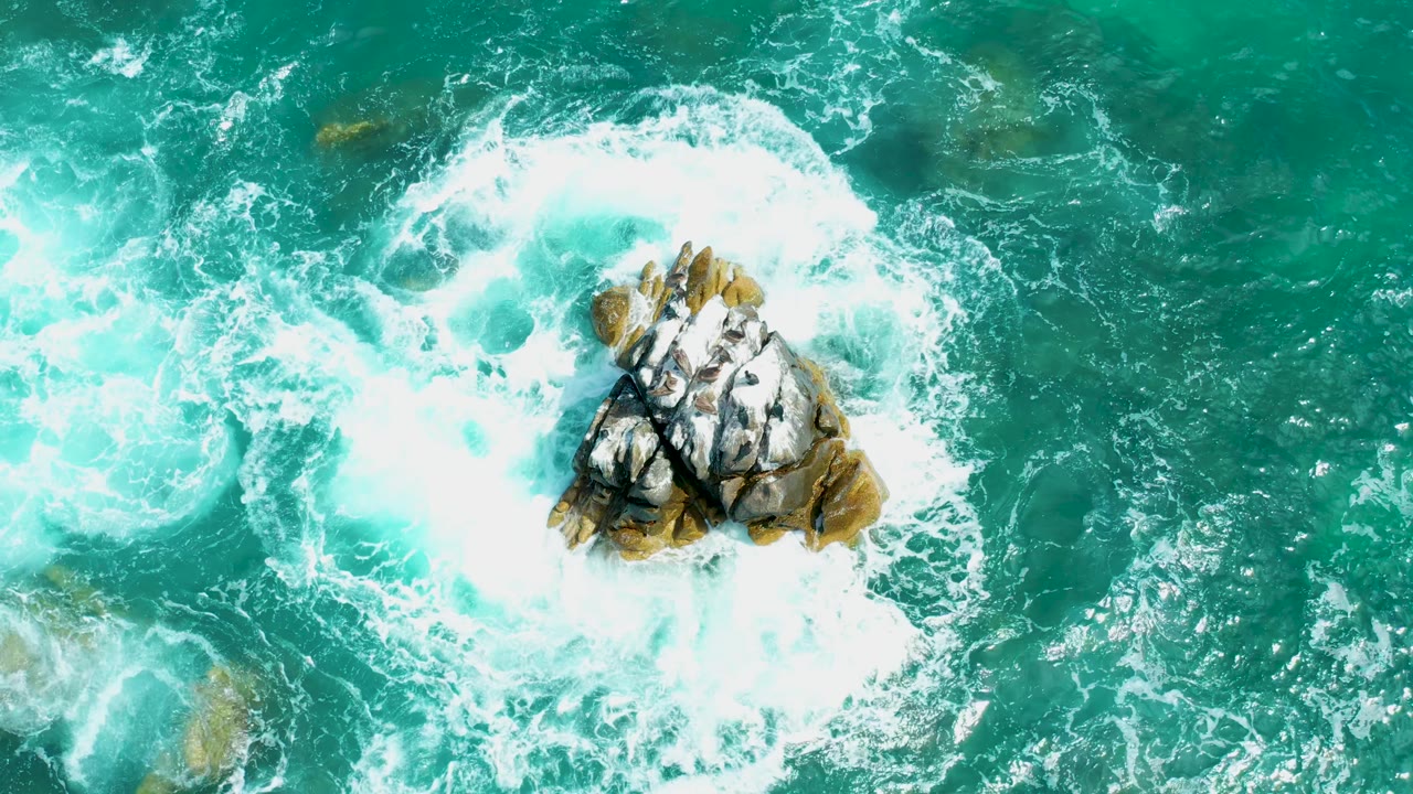 Top-down view at Marquis Los Cabos, Mexico, with a rounded white wave wash curling around the rocky outcrop and a patch of calmer turquoise water above it, with no visible people.