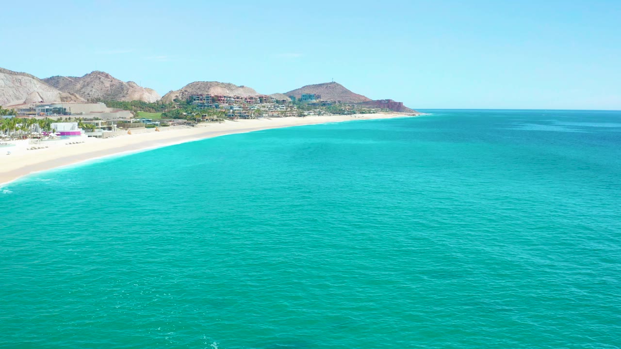 Aerial frame over turquoise ocean at Marquis Los Cabos, with the pale beach curving along the left side, shoreline buildings beyond it, and no visible people.