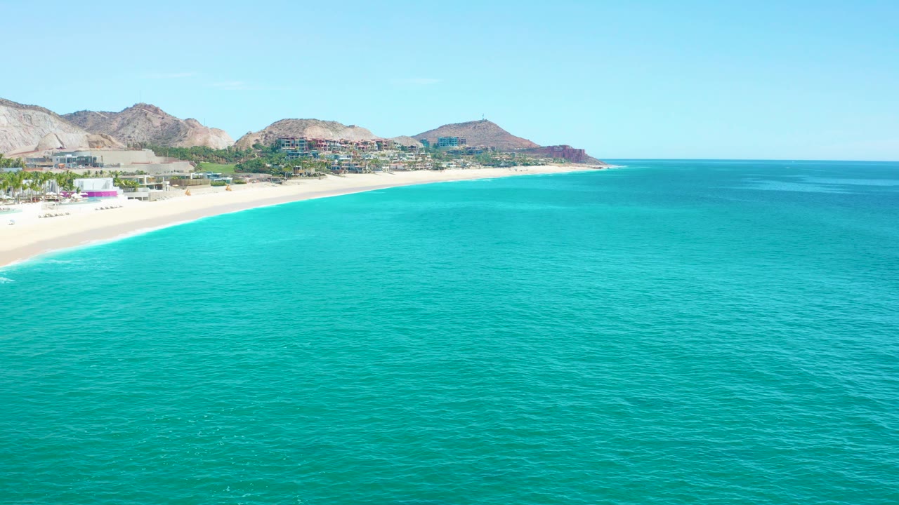 Turquoise sea with beach and coastal development below low desert mountains.
