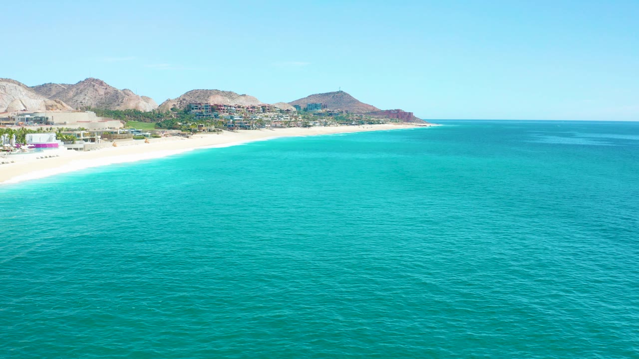 Opening frame of Marquis Los Cabos, Mexico, showing turquoise ocean in the foreground, a white sand shoreline at left, coastal buildings, dry mountains, clear blue sky, and no visible people.
