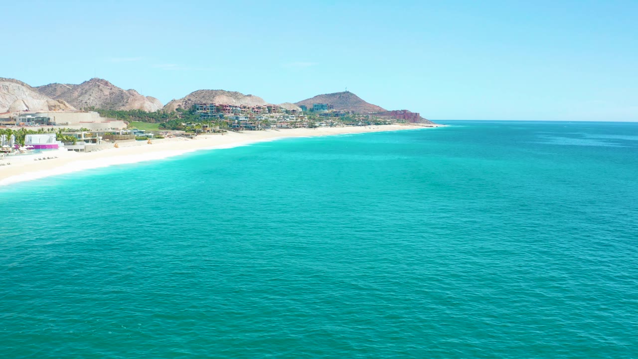 A bright aerial frame over Marquis Los Cabos shows open turquoise sea, a narrow white shoreline curving left to right, low buildings near the beach, mountains behind, and no visible people.