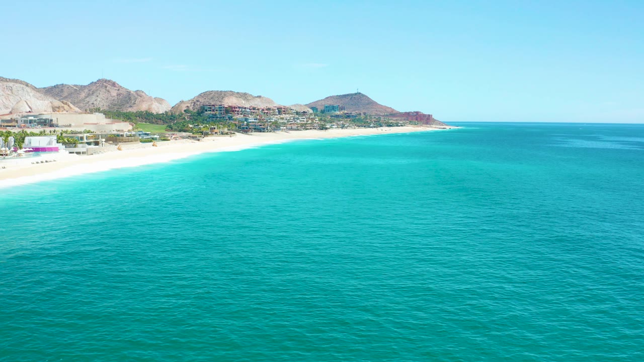 White beach and turquoise ocean with distant buildings under a clear sky.
