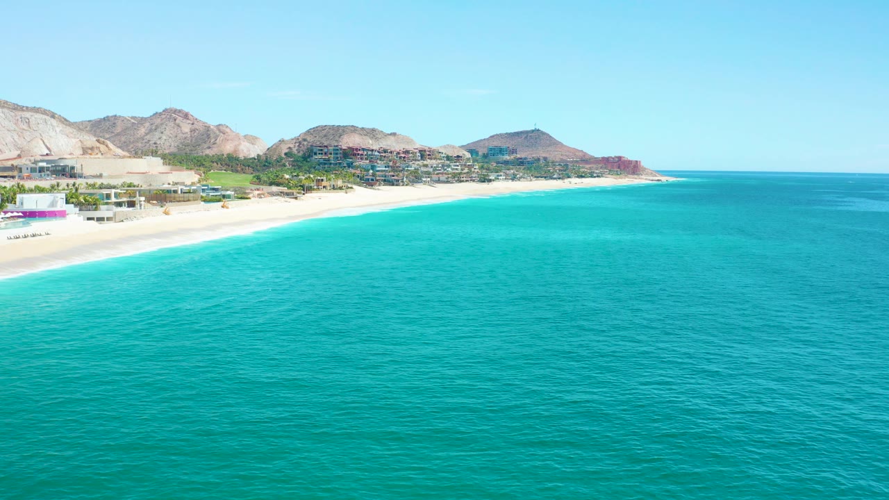 Wide aerial frame at Marquis Los Cabos, Mexico, with turquoise water in the foreground, pale sand at left, coastal buildings, mountains, clear sky, and no visible people.