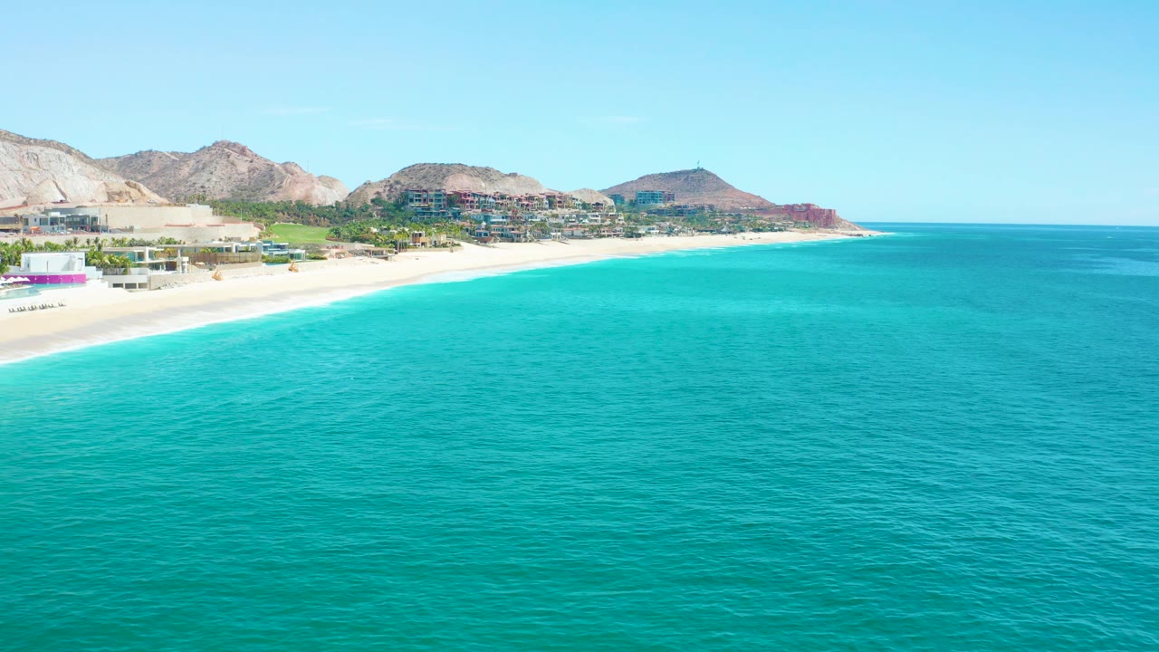 Aerial view over bright turquoise water at Marquis Los Cabos, Mexico, with the sandy shoreline running diagonally toward coastal buildings and mountains, with no visible people.