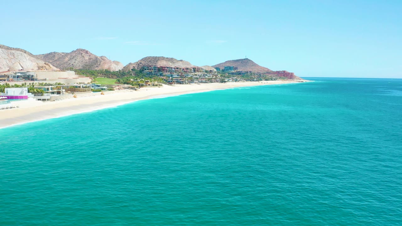 Wide aerial frame of Marquis Los Cabos, Mexico, showing textured turquoise sea across the foreground, a pale beach at left, resort-area buildings, rocky mountains, and no visible people.