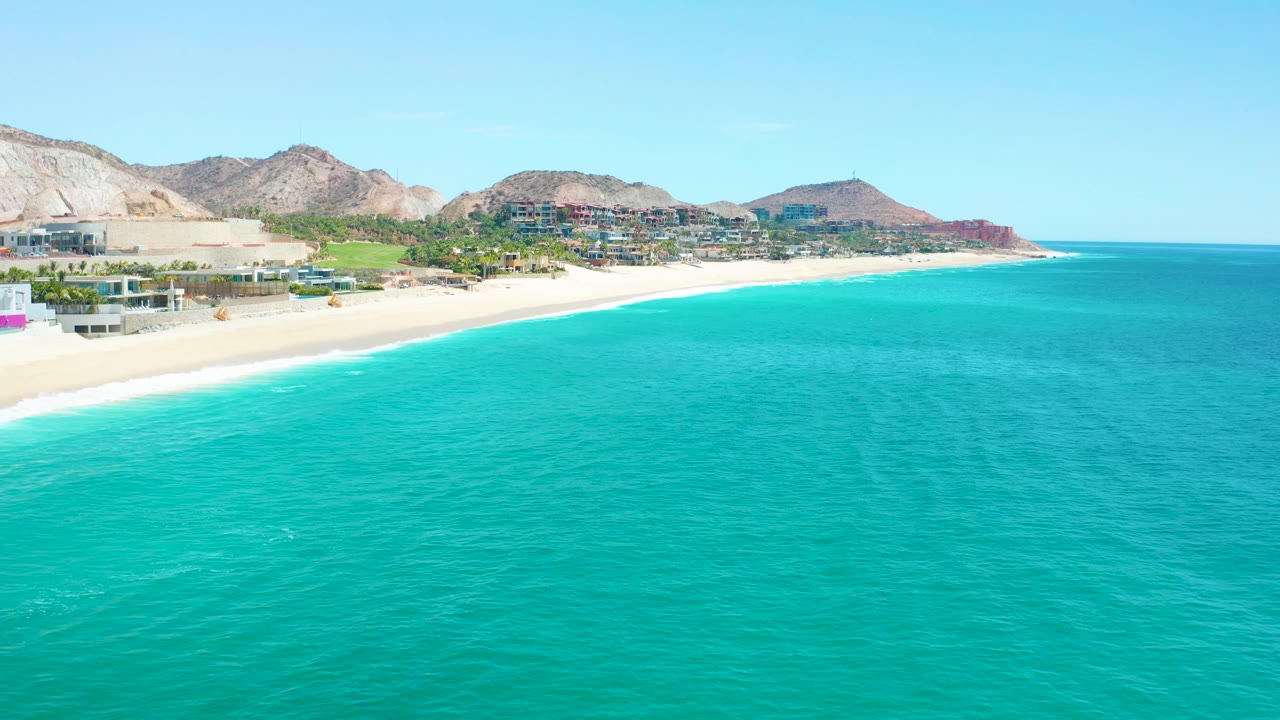 Aerial frame over turquoise water looking toward the Marquis Los Cabos beach, with shoreline buildings at left, mountains behind them, and no visible people.