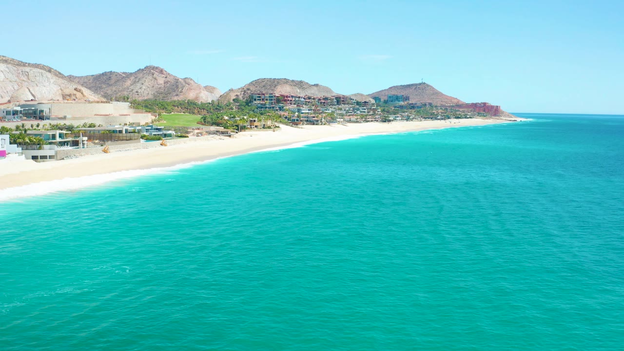 The Marquis Los Cabos coast appears from an elevated angle with broad turquoise ocean, a narrow white surf edge, beach buildings, dry hills, and no visible people.