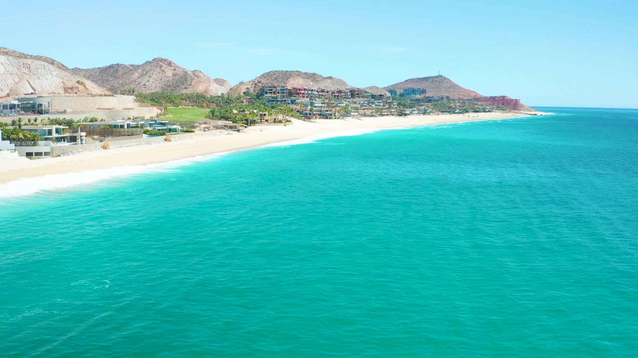 A wide coastal frame shows turquoise water below a sandy Marquis Los Cabos beach, low buildings near the shore, tan mountains in the distance, and no visible people.
