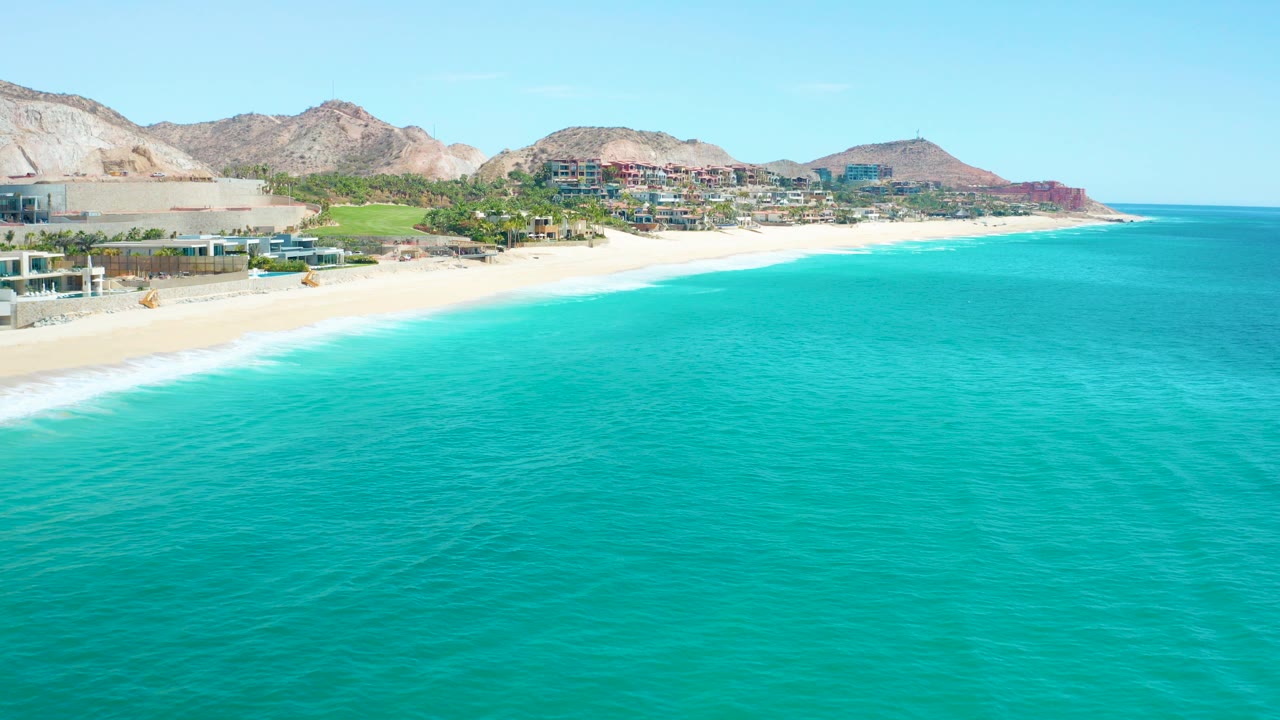 Opening aerial frame over Marquis Los Cabos, Mexico, with turquoise water in the foreground, a pale sandy beach at left, coastal buildings and rocky hills beyond, and no visible people.