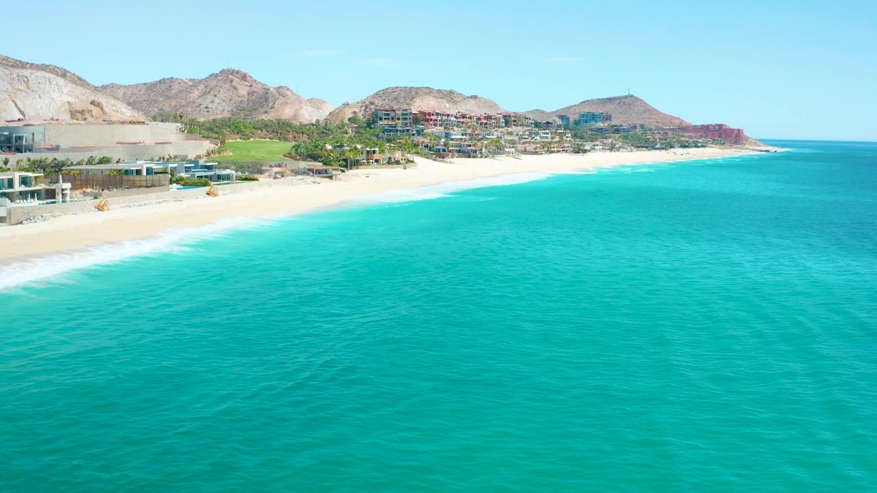 Aerial view at 0:01 over Marquis Los Cabos, Mexico, showing broad turquoise water, a curving sand beach, distant coastal buildings, rocky hills, and no visible people.