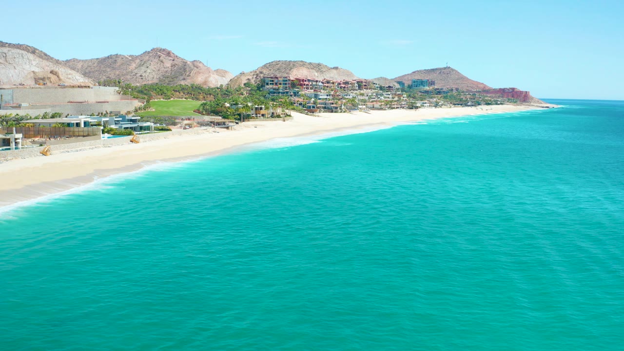Aerial beach view with turquoise sea in front and coastal buildings below rocky hills.