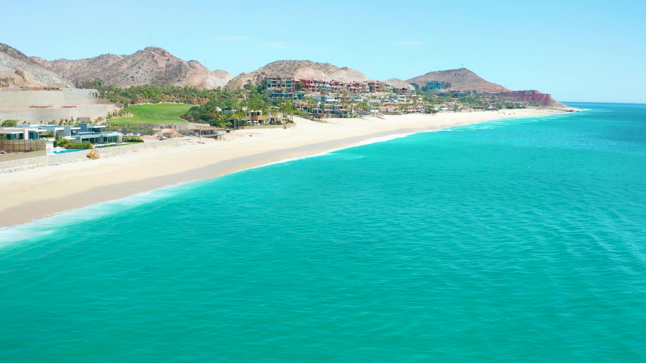 Wide aerial view of turquoise water meeting a sandy beach at Marquis Los Cabos, Mexico, with coastal buildings and rocky mountains in the background and no visible people.