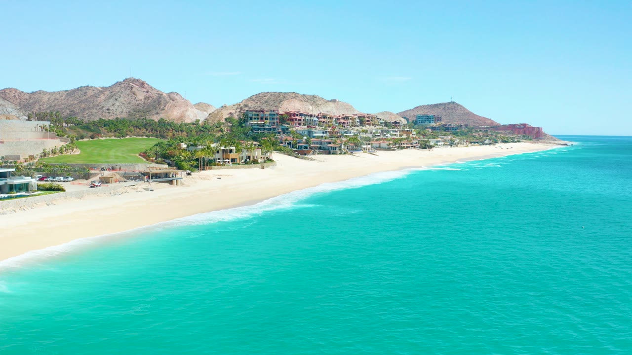 Wide elevated view of Marquis Los Cabos coastline with turquoise water filling the foreground, pale sand on the left, hillside buildings, rocky mountains, clear sky, and no visible people.