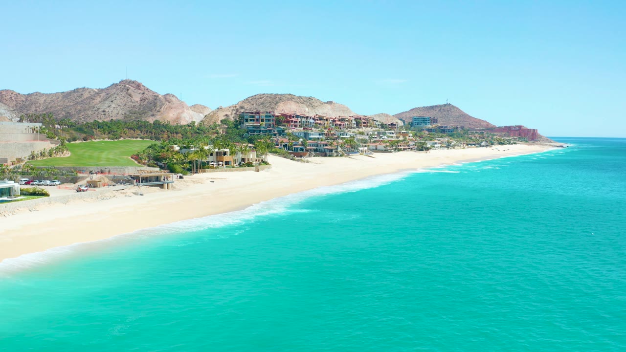 Elevated Marquis Los Cabos coast view with the waterline angled across the frame, blue-green ocean in front, sand and hillside buildings behind it, rocky mountains, clear sky, and no visible people.