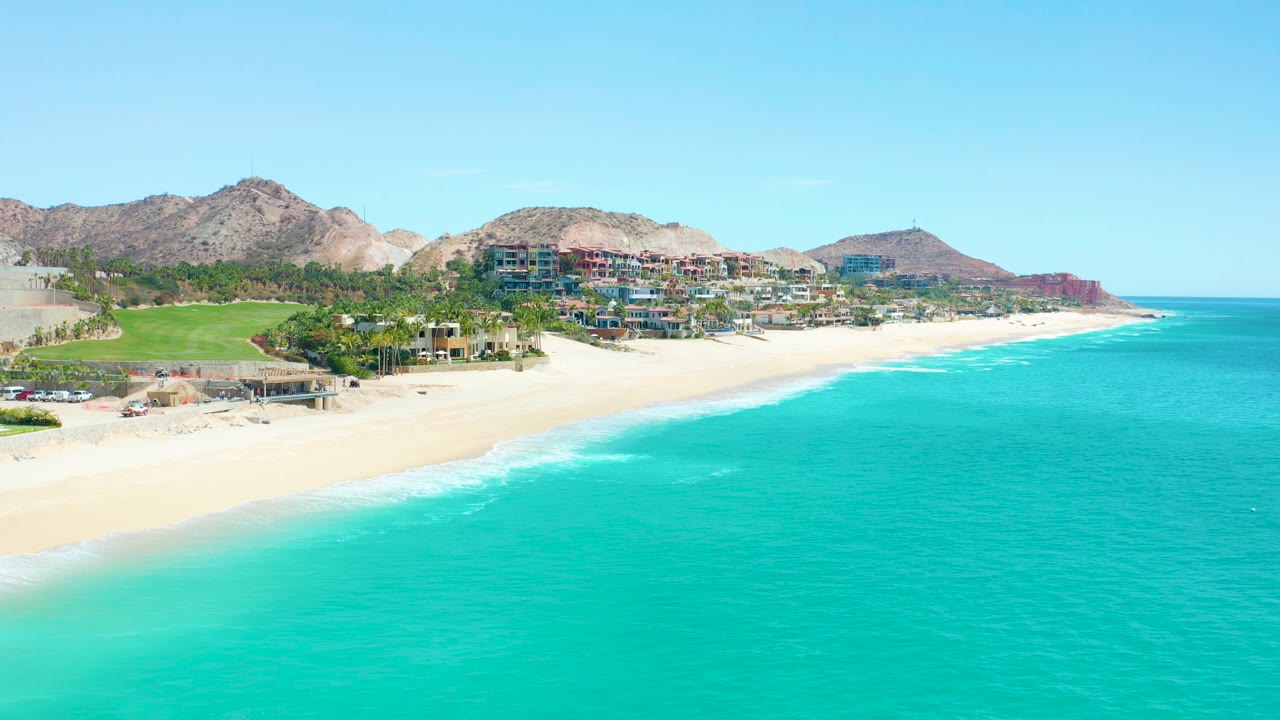 Beach and turquoise sea with green grounds, buildings, and rocky hills behind.